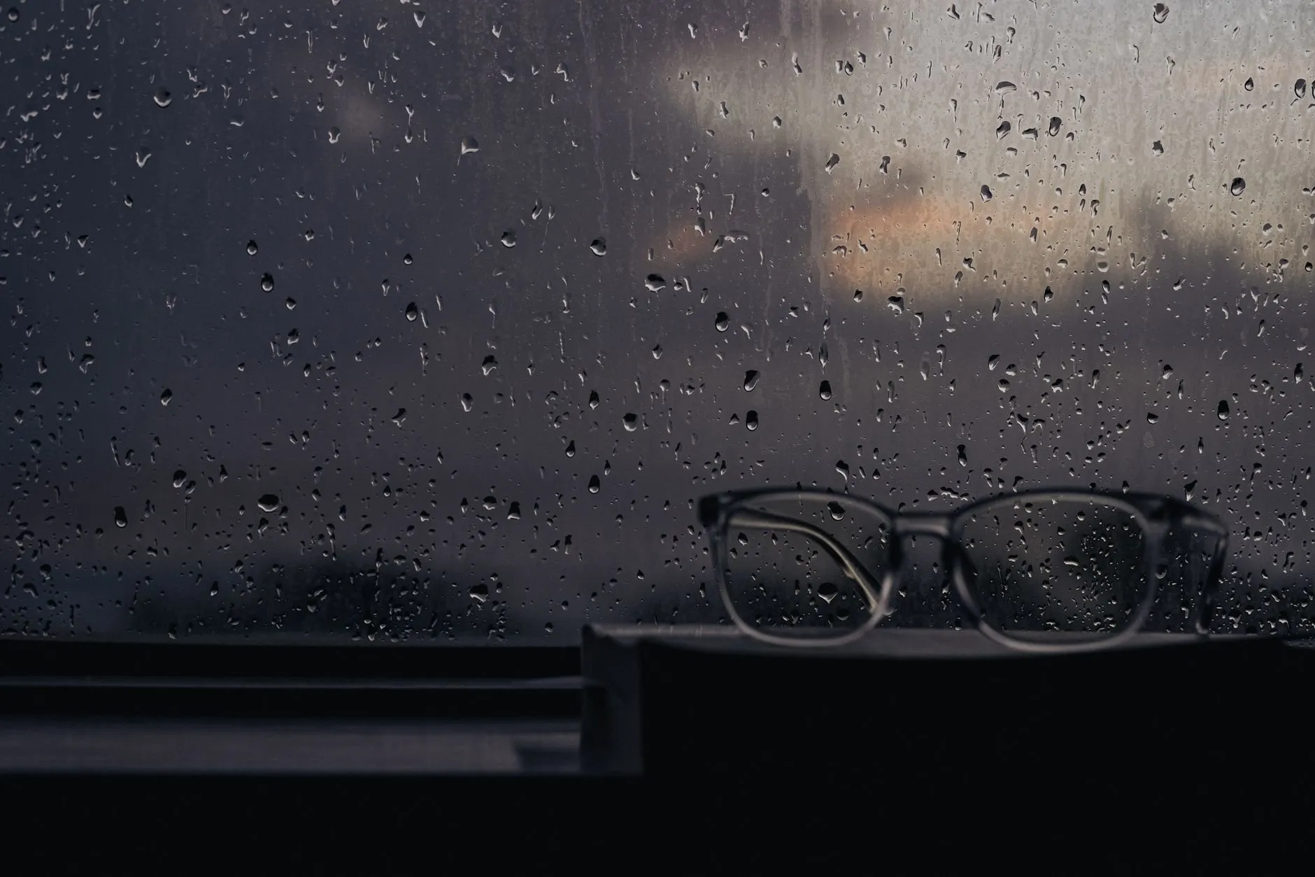 Reading glasses on a windowsill behind a rain-streaked pane at dusk