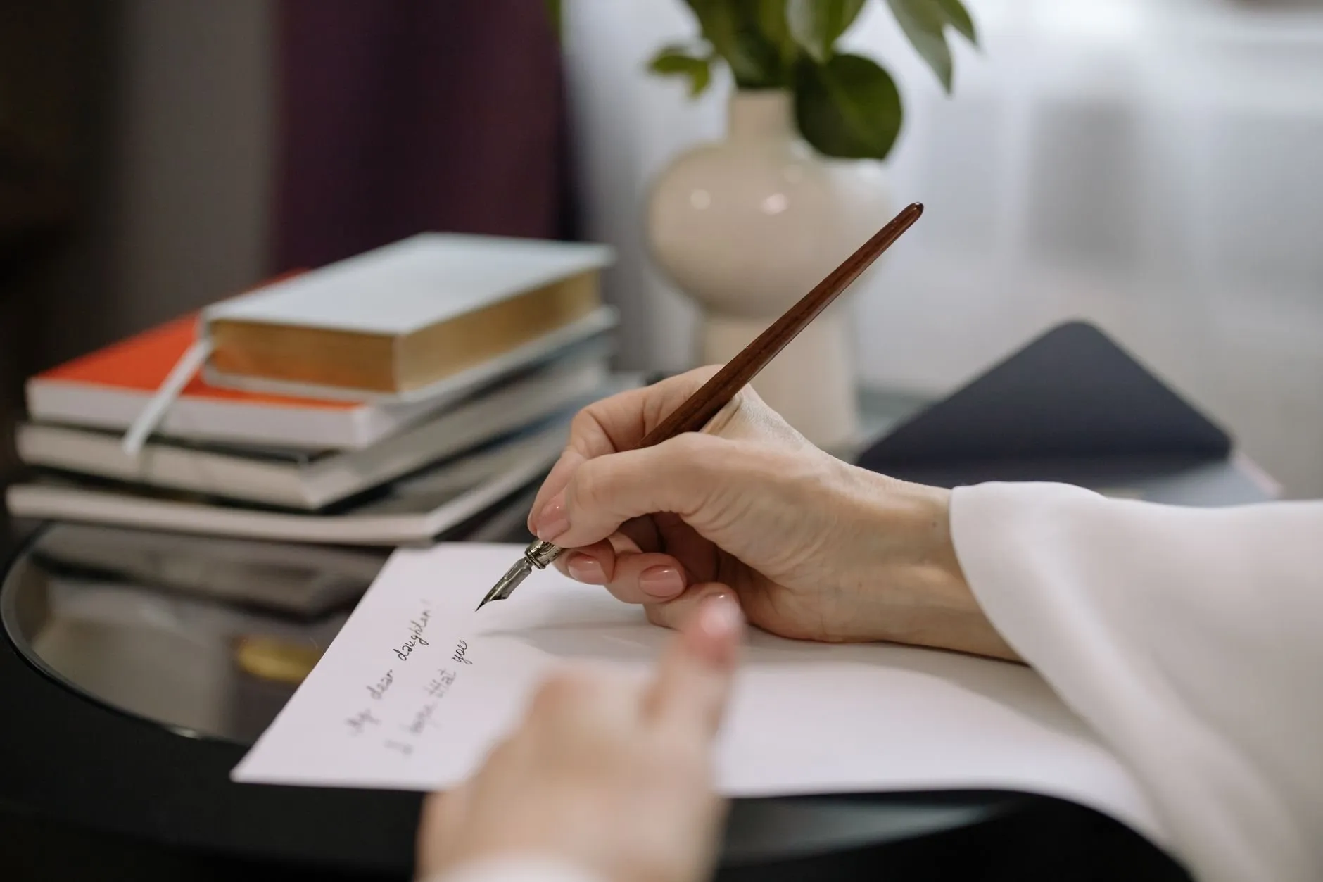 A writer's hands with a dip pen over a handwritten letter, books stacked behind