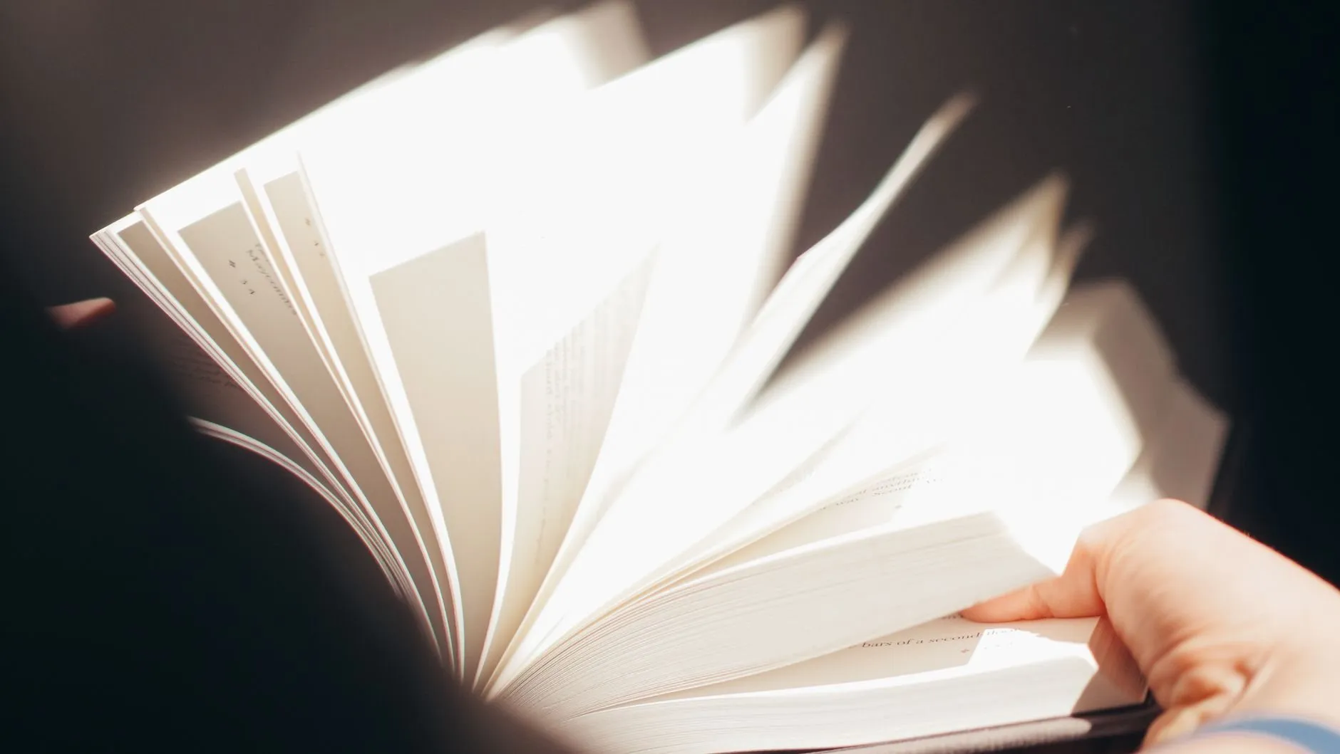 Hands fanning the pages of a book in a beam of window light against a dark background