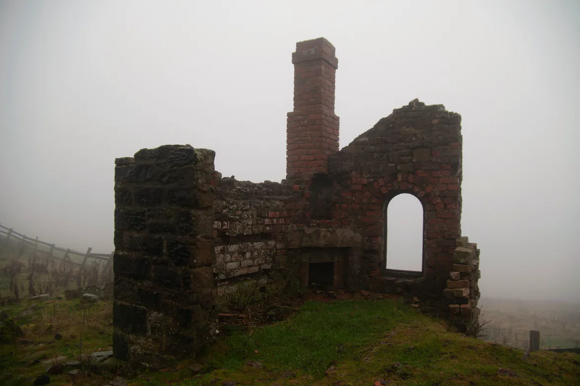 The ruin of a stone engine house standing in mist, its chimney still up.